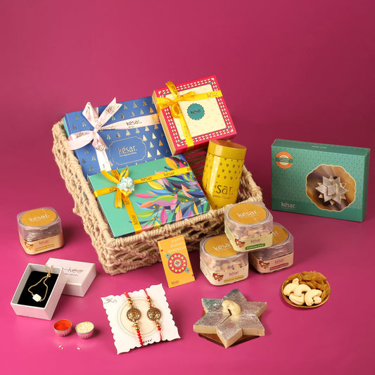 Assortment of Rakhi gift hampers displayed on a pink background. The centerpiece is a woven basket filled with various colorful and beautifully wrapped gift boxes labeled 'Kesar,' each tied with decorative ribbons. Surrounding the basket are more gift boxes, jars of dry fruits, traditional Indian sweets, and a pair of intricate Rakhis. Additional items include a jewelry box with a necklace, small containers of kumkum and rice, and a card reading 'Happy Raksha Bandhan.
