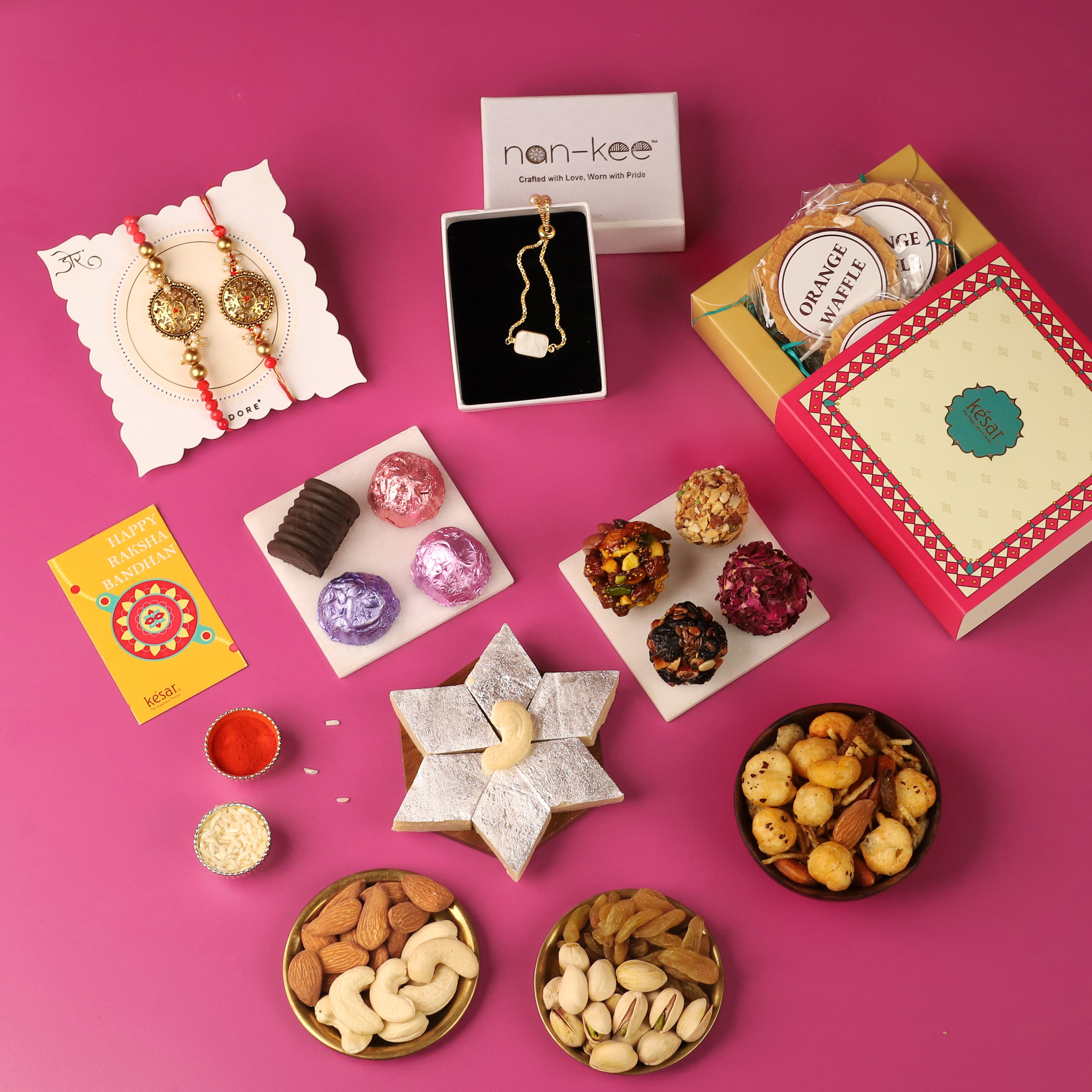 Flat lay of Rakhi gift items on a pink background, featuring a pair of intricately designed Rakhis on a card, a jewelry box with a 'nan-kee' necklace, assorted traditional Indian sweets, a box of orange wafers, and a 'Happy Raksha Bandhan' card. Additional items include small containers of kumkum and rice, dry fruit platters with almonds, cashews, raisins, and pistachios, and a variety of snacks. The image showcases a festive and diverse selection of gifts for Raksha Bandhan.