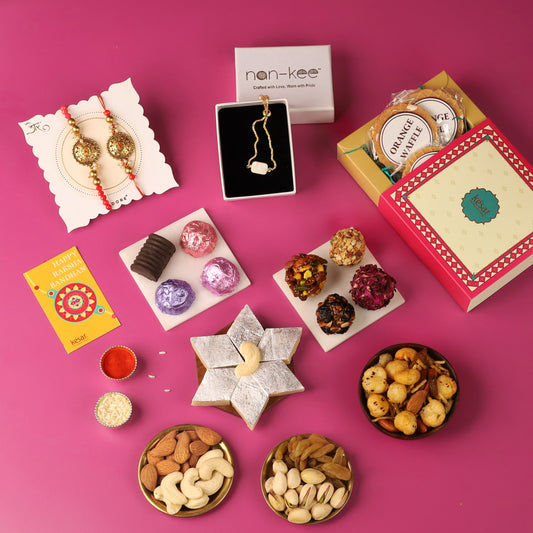 Flat lay of Rakhi gift items on a pink background, featuring a pair of intricately designed Rakhis on a card, a jewelry box with a 'nan-kee' necklace, assorted traditional Indian sweets, a box of orange wafers, and a 'Happy Raksha Bandhan' card. Additional items include small containers of kumkum and rice, dry fruit platters with almonds, cashews, raisins, and pistachios, and a variety of snacks. The image showcases a festive and diverse selection of gifts for Raksha Bandhan.